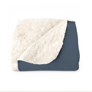 May include: A folded throw blanket featuring a solid dark slate blue exterior and a thick, cream-coloured faux sheepskin interior lining. The blanket is partially folded to reveal both the smooth outer fabric and the textured, plush inner fleece.