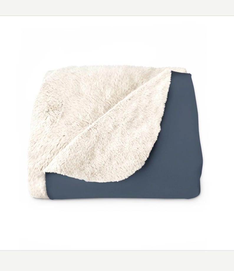 May include: A folded throw blanket featuring a solid dark slate blue exterior and a thick, cream-coloured faux sheepskin interior lining. The blanket is partially folded to reveal both the smooth outer fabric and the textured, plush inner fleece.