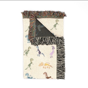 May include: A woven throw blanket with a cream-coloured base featuring a repeating pattern of small, multi-coloured dinosaurs in shades of green, blue, purple, yellow, and brown. The blanket has a dark, solid-coloured reverse side and fringed edges.