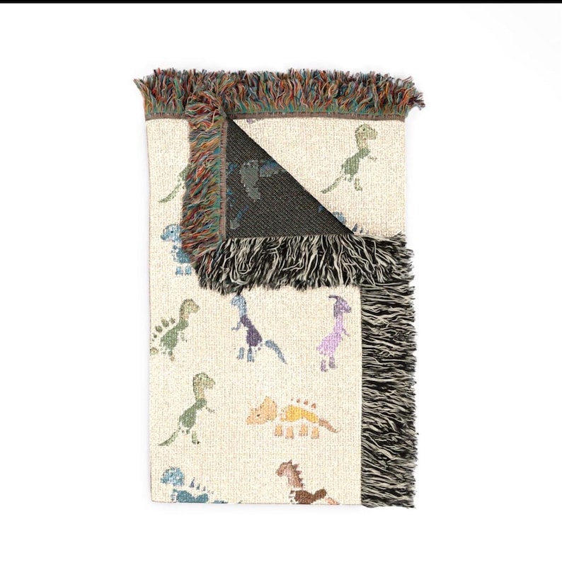 May include: A woven throw blanket with a cream-coloured base featuring a repeating pattern of small, multi-coloured dinosaurs in shades of green, blue, purple, yellow, and brown. The blanket has a dark, solid-coloured reverse side and fringed edges.