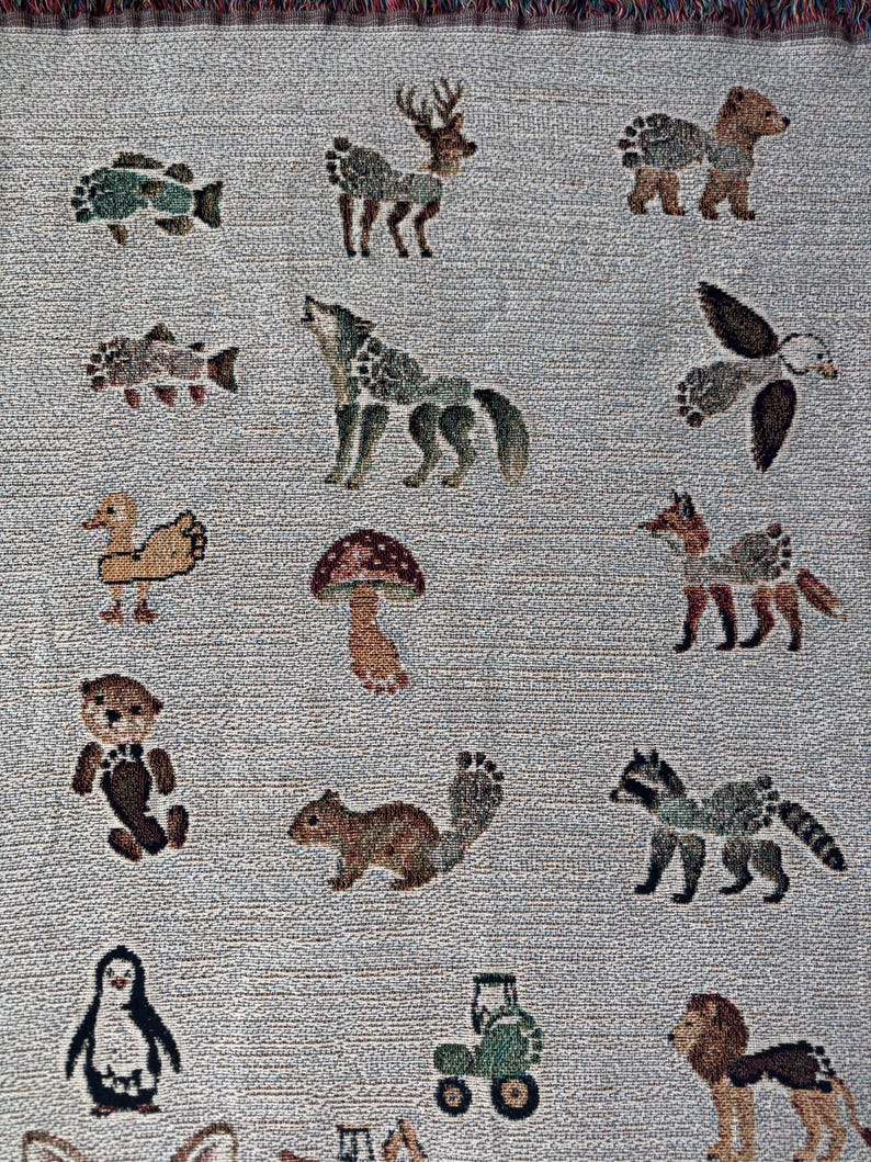 May include: A woven throw blanket featuring a light grey background with a repeating pattern of various animals including a deer, wolf, bear, fox, raccoon, squirrel, otter, penguin, lion, duck, and fish, alongside a mushroom and a small green tractor.