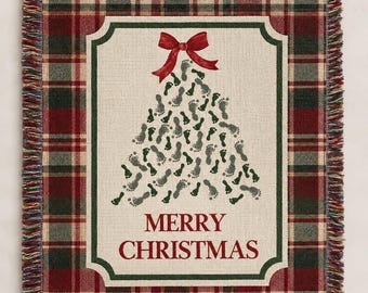 May include: A woven throw blanket featuring a red, green, and beige plaid border with fringe edges. The centre panel is cream-coloured with a Christmas tree shape made of small footprints, a red bow at the top, and the text Merry Christmas.