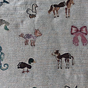 May include: A light grey woven fabric featuring a repeating pattern of small, colourful embroidered motifs including a white duck, a brown horse, a pink bow, a small fawn, a dark duck, a spotted foal, and a stylised angel figure in black.