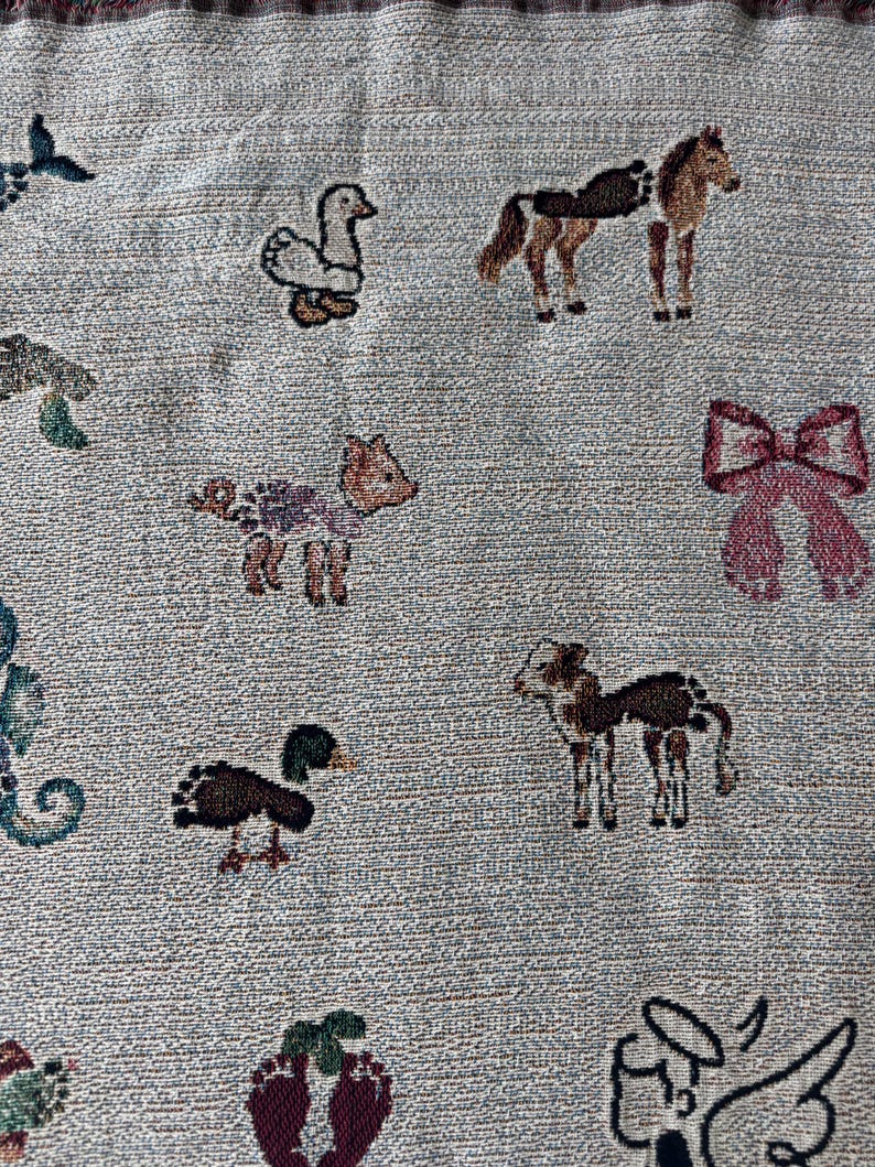 May include: A light grey woven fabric featuring a repeating pattern of small, colourful embroidered motifs including a white duck, a brown horse, a pink bow, a small fawn, a dark duck, a spotted foal, and a stylised angel figure in black.