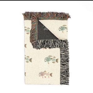 May include: A woven throw blanket with a cream-coloured base featuring a repeating pattern of multi-coloured fish. The blanket has a folded corner revealing a dark interior and is finished with decorative multi-coloured fringe along the edges.