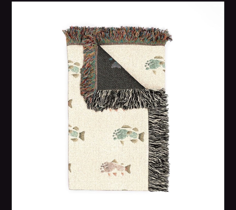 May include: A woven throw blanket with a cream-coloured base featuring a repeating pattern of multi-coloured fish. The blanket has a folded corner revealing a dark interior and is finished with decorative multi-coloured fringe along the edges.