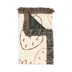 May include: A woven throw blanket featuring a cream-coloured base with a repeating pattern of outlined strawberries in a reddish-brown hue. The blanket has a folded corner revealing a dark interior with the partial text OTTE visible in green.