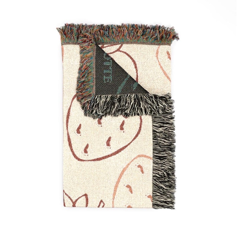 May include: A woven throw blanket featuring a cream-coloured base with a repeating pattern of outlined strawberries in a reddish-brown hue. The blanket has a folded corner revealing a dark interior with the partial text OTTE visible in green.