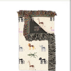 May include: A cream-coloured woven throw blanket featuring a repeating pattern of various animals including giraffes, lions, elephants, and crocodiles. The blanket has a folded corner revealing a dark interior and multi-coloured fringed edges.