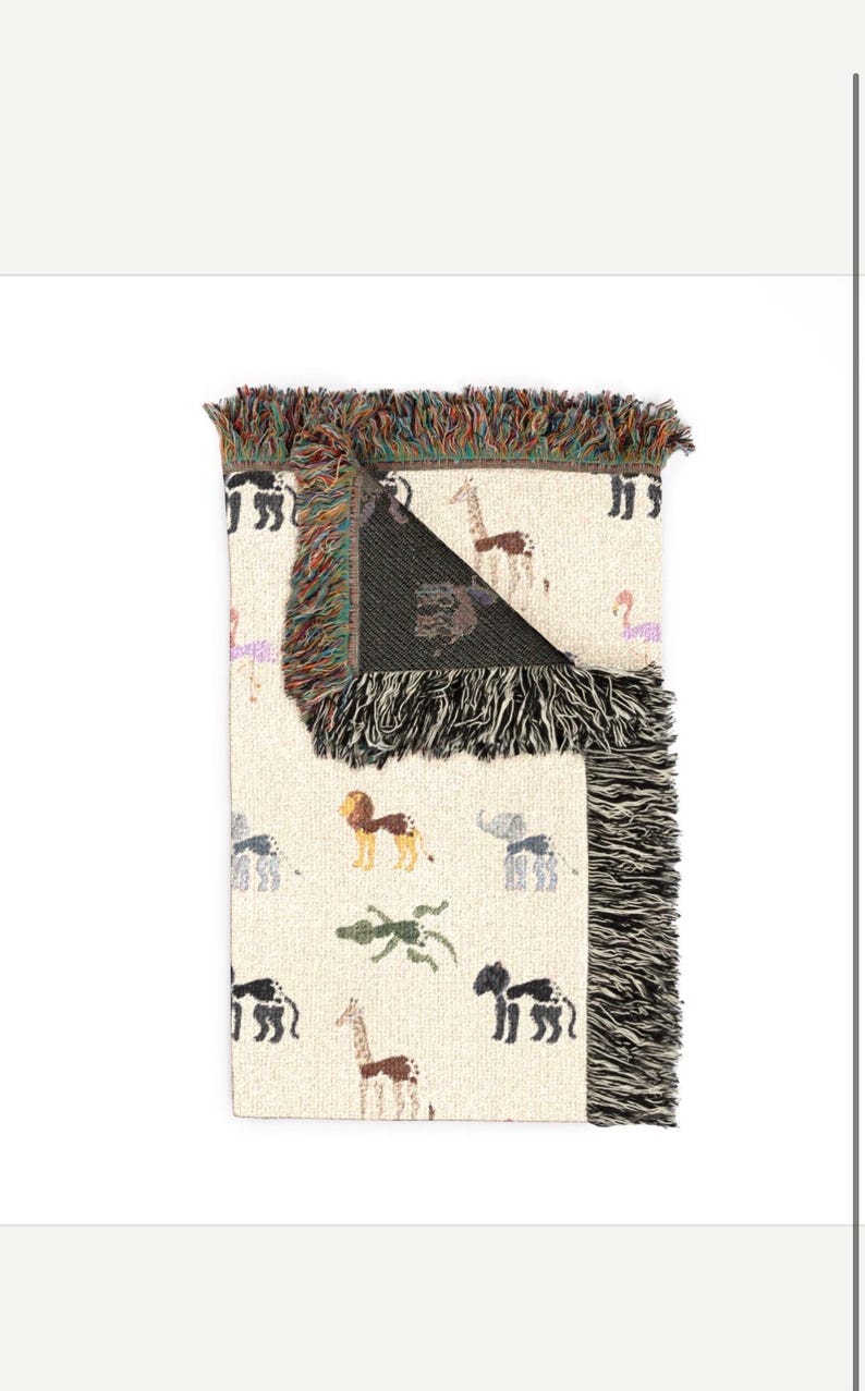 May include: A cream-coloured woven throw blanket featuring a repeating pattern of various animals including giraffes, lions, elephants, and crocodiles. The blanket has a folded corner revealing a dark interior and multi-coloured fringed edges.