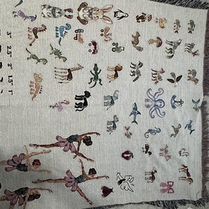 May include: A woven throw blanket featuring a light beige background with a variety of small, colourful embroidered motifs including animals, dancers, and vehicles. A printed scale shows measurements from 1 inch to 3 inches along the left side.