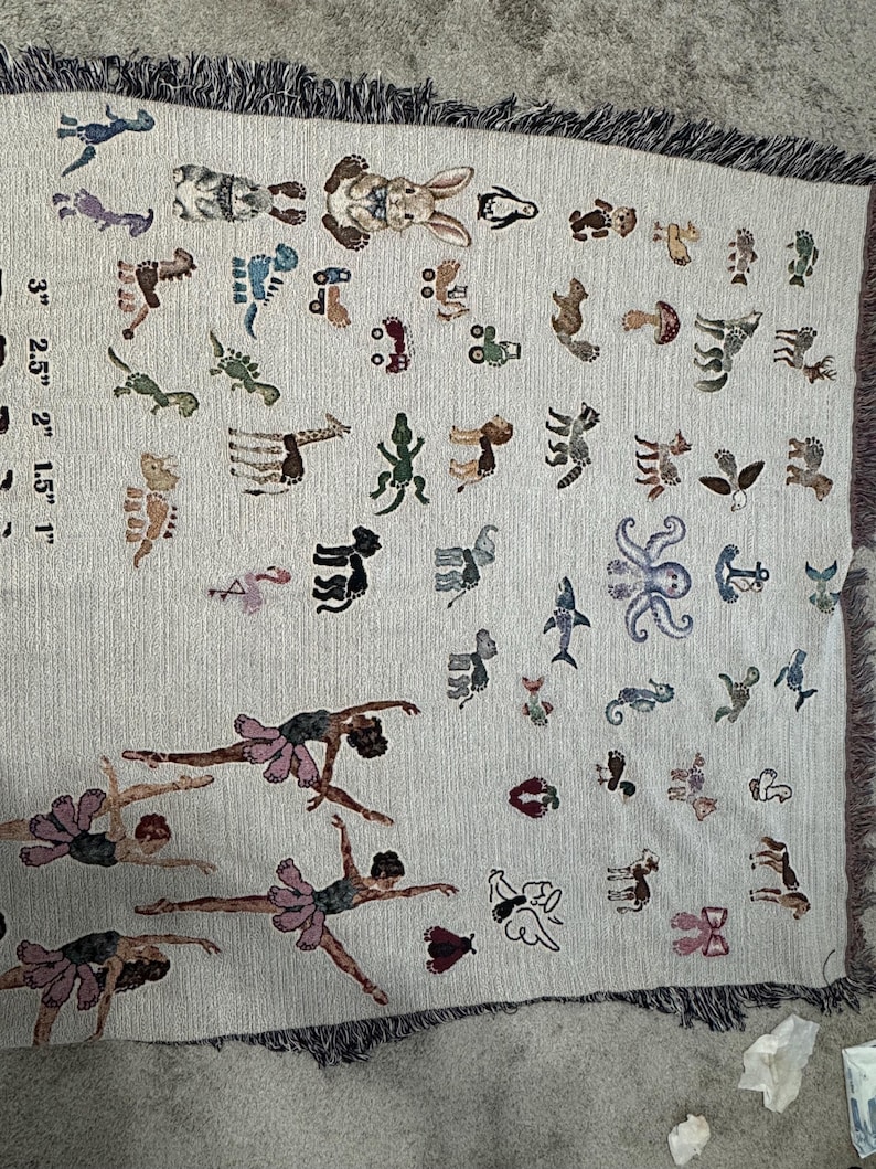 May include: A woven throw blanket featuring a light beige background with a variety of small, colourful embroidered motifs including animals, dancers, and vehicles. A printed scale shows measurements from 1 inch to 3 inches along the left side.