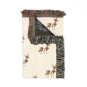 May include: A woven throw blanket with a cream-colored base featuring a repeating pattern of brown deer and paw prints. The blanket has a folded corner revealing a dark reverse side and is finished with multicolored fringed edges on all sides.