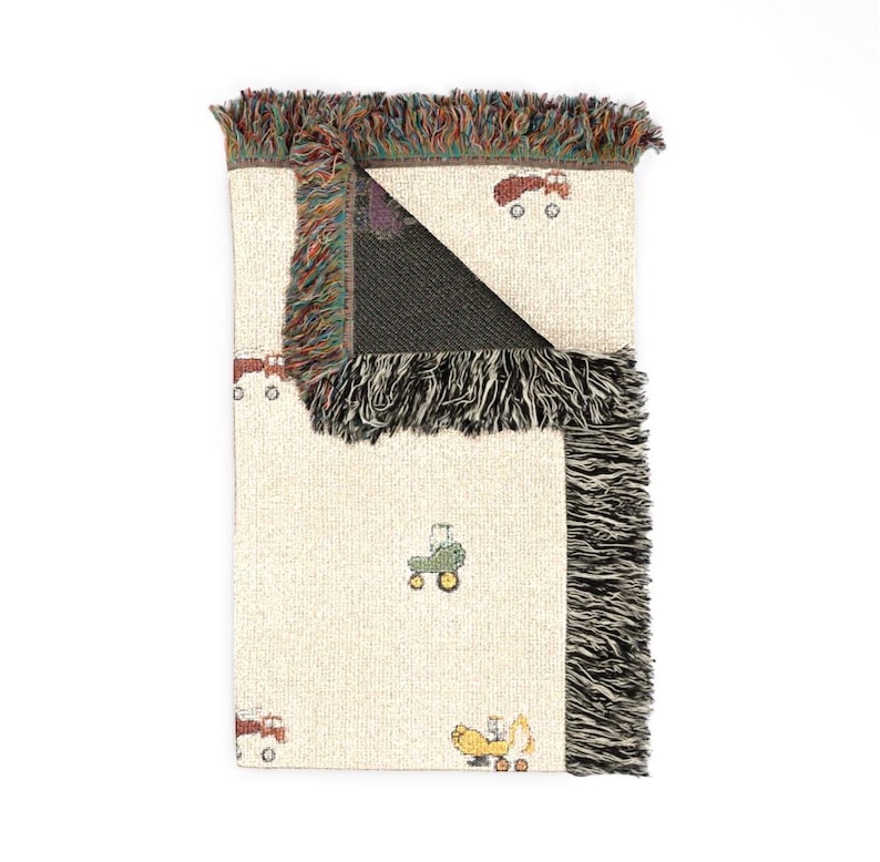 May include: A woven throw blanket with a cream-coloured base featuring a repeating pattern of small, colourful tractors in red, green, and yellow. The blanket has a textured, fringed edge with multi-coloured threads along the top and side borders.