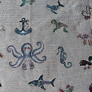 May include: A woven throw blanket featuring a light grey background with a repeating pattern of various sea creatures including a blue octopus, a shark, a seahorse, a turtle, a whale, and a fish, alongside a blue anchor and a white duck.