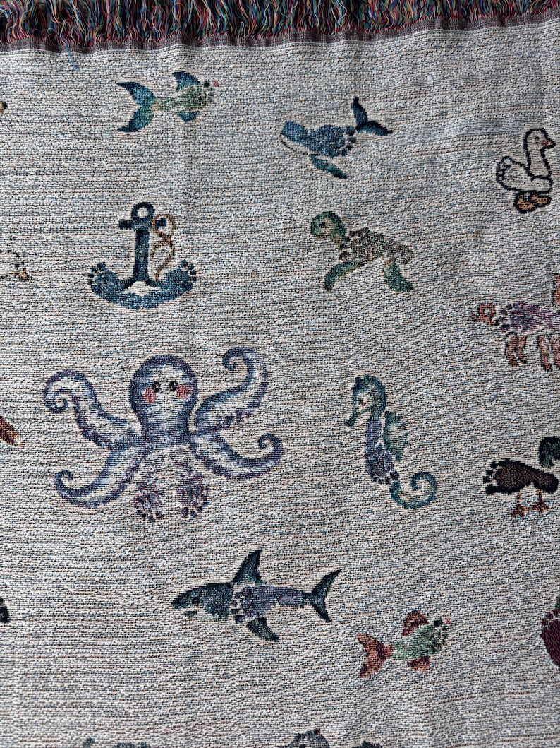 May include: A woven throw blanket featuring a light grey background with a repeating pattern of various sea creatures including a blue octopus, a shark, a seahorse, a turtle, a whale, and a fish, alongside a blue anchor and a white duck.