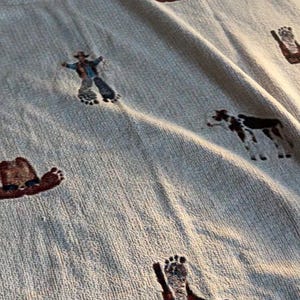 May include: A light beige woven throw blanket featuring a repeating pattern of embroidered motifs including brown cowboy boots with footprints, brown cowboy hats, a standing brown horse, a standing cow, and a figure wearing a cowboy hat.