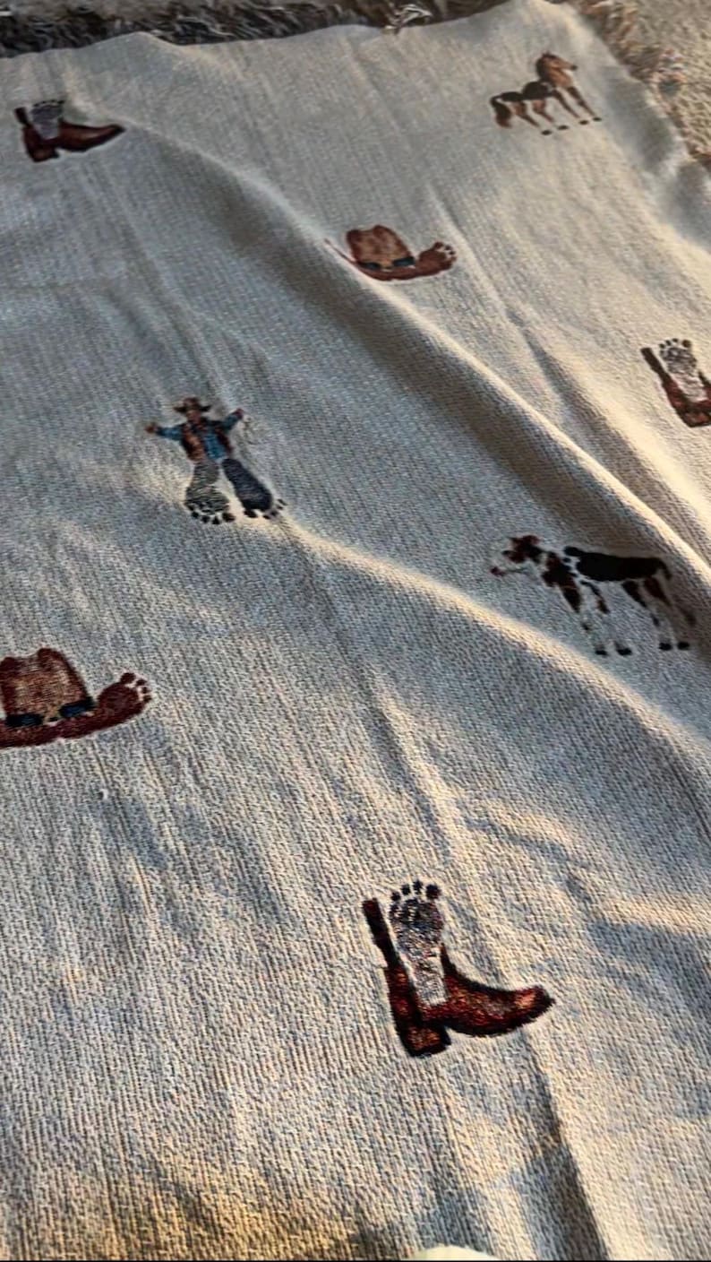 May include: A light beige woven throw blanket featuring a repeating pattern of embroidered motifs including brown cowboy boots with footprints, brown cowboy hats, a standing brown horse, a standing cow, and a figure wearing a cowboy hat.
