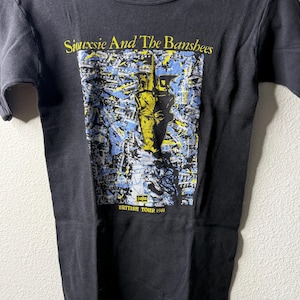 Op de afbeelding: Zwart T-shirt met de bandnaam "Siouxsie And The Banshees" in gele letters. De grafische print eronder toont gele en blauwe elementen, met de tekst "British Tour 1981".