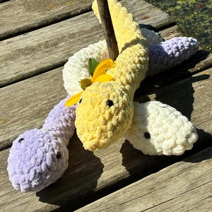May include: Crocheted snake toys in yellow, white, and lavender. The plush snakes have black button eyes and are arranged on a wooden surface. A yellow flower is placed near the yellow snake's head.