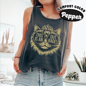 May include: A dark grey sleeveless tank top featuring a gold graphic of a cat wearing a crown and round sunglasses. The text inside the glasses reads THIS IS AN OUTRAGE. The shirt is labelled as Comfort Colour in the colour Pepper.