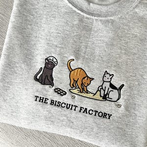 May include: Light gray sweatshirt with embroidered design of three cats and the text "THE BISCUIT FACTORY". The cats are black, orange, and white, with one wearing a chef's hat. The sweatshirt is folded on a light gray surface.