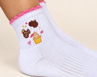 May include: White ribbed ankle socks featuring a bright pink scalloped lettuce edge trim at the cuff. The socks are decorated with small embroidered icons including a brown chocolate-covered treat, a yellow soft-serve ice cream, and pink bows.