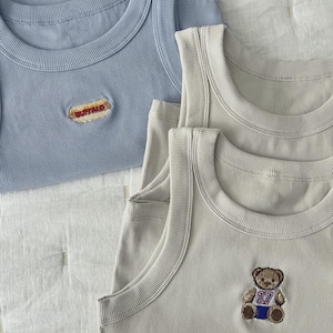 May include: Close-up of several ribbed tank tops in light blue and beige. One light blue tank top has a "BUFFALO" patch. A beige tank top has a teddy bear patch with the number "17" on it. The tank tops are stacked on a white, textured surface.