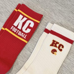 Pode incluir: Dois pares de meias esportivas. Um par &eacute; vermelho com texto branco "KC FOOTBALL" e listras amarelas. O outro par &eacute; branco sujo com listras vermelhas e um logotipo "KC" com um capacete de futebol americano.