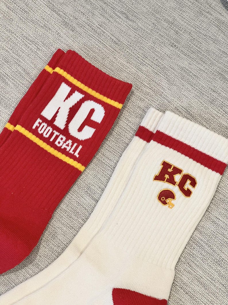 Pode incluir: Dois pares de meias esportivas. Um par &eacute; vermelho com texto branco "KC FOOTBALL" e listras amarelas. O outro par &eacute; branco sujo com listras vermelhas e um logotipo "KC" com um capacete de futebol americano.