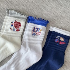 May include: Three pairs of white and navy blue crew socks with ruffled cuffs. Each sock features embroidered designs: a heart with "Buffalo Football Club", the number "17" with a buffalo, and football-themed graphics with "Game Day".