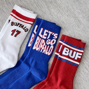 May include: Three crew socks in white, blue, and red. The white sock features red stripes and the text BUFFALO 17. The blue sock displays the text LET'S GO BUFFALO in white and red. The red sock has blue and white stripes with the text BUF.