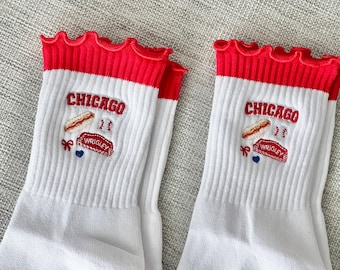 Pode incluir: Um par de meias brancas com bordas vermelhas com babados. As meias apresentam bordados de "CHICAGO", um cachorro-quente e "WRIGLEY". O design utiliza vermelho, branco e azul, possivelmente em referência a Chicago. Ideal para fãs.