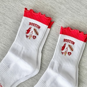 May include: Pair of white socks with red ruffled tops. Each sock features the word "BOSTON" above embroidered images of a lobster, hot dog, ice cream, and a heart with a "B".