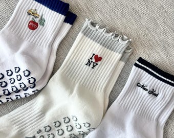 May include: Three pairs of white socks with ribbed texture and various New York City-themed designs. One pair features a taxi and apple embroidery, another has "I NY" with a heart, and the third says "New York".