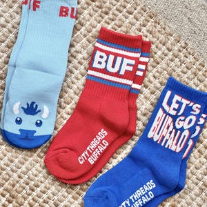 May include: Three pairs of crew socks in light blue, red, and blue. The light blue socks have a buffalo graphic and the word "BUF". The red socks have "BUF" and stripes. The blue socks say "LET'S GO BUFFALO". All socks say "CITY THREADS BUFFALO".