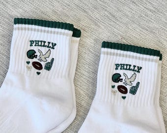 May include: A pair of white crew socks with a dark green ribbed cuff. The socks feature an embroidered design with the text PHILLY in dark green, accompanied by a dark green football helmet, a white eagle, a brown football, and a green foam finger.