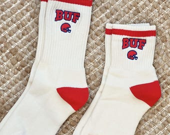 May include: A pair of white crew socks with red accents. The socks have red toes and a red band at the top. The word "BUF" is embroidered in red and blue, along with a blue football helmet.