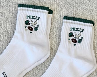 May include: Pair of white ankle socks with green and tan striped trim. Each sock features the word "PHILLY" above embroidered football-themed graphics, including a helmet, football, and a hand making a "number one" gesture. The word "THREADS" is printed on the side.