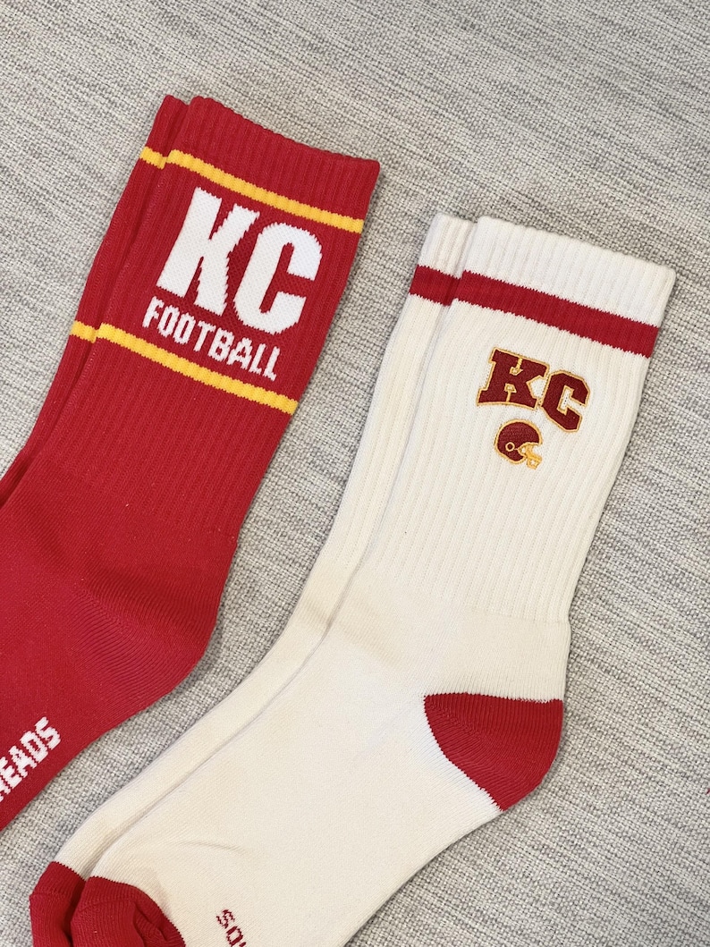 Pode incluir: Dois pares de meias esportivas. Um par &eacute; vermelho com texto branco "KC FOOTBALL" e listras amarelas. O outro par &eacute; branco com detalhes vermelhos e um logotipo "KC" com um capacete de futebol. Ambos os pares t&ecirc;m textura canelada.
