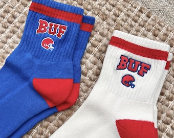May include: Two pairs of crew socks. One pair is blue with red accents and the letters "BUF" in white with a red outline and a red football helmet graphic. The other pair is white with red accents and the same "BUF" design.