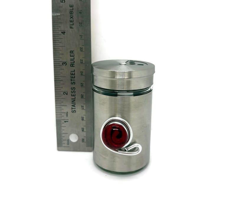 May include: A stainless steel spice jar with a glass insert and a red glass swirl accent. The jar has a screw-on lid with a small hole for dispensing spices.
