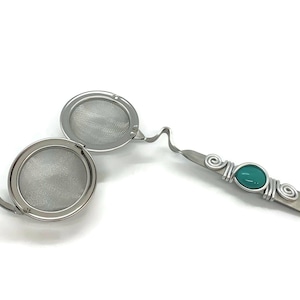 May include: A stainless steel tea infuser with a teal colored gem on the handle. The infuser has two round mesh baskets that can be used to steep loose leaf tea.