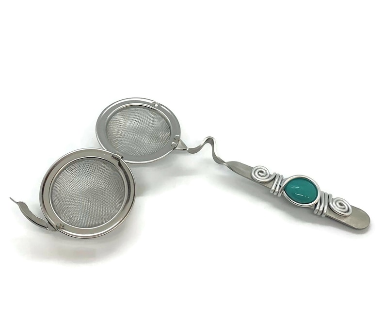 May include: A stainless steel tea infuser with a teal colored gem on the handle. The infuser has two round mesh baskets that can be used to steep loose leaf tea.