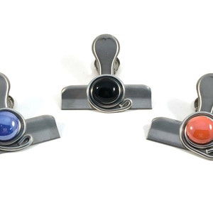 May include: Three metal binder clips with colored accents. One clip has a blue orb, another a black orb, and the third a coral orb. Each clip has a silver-colored body and a decorative wire swirl.