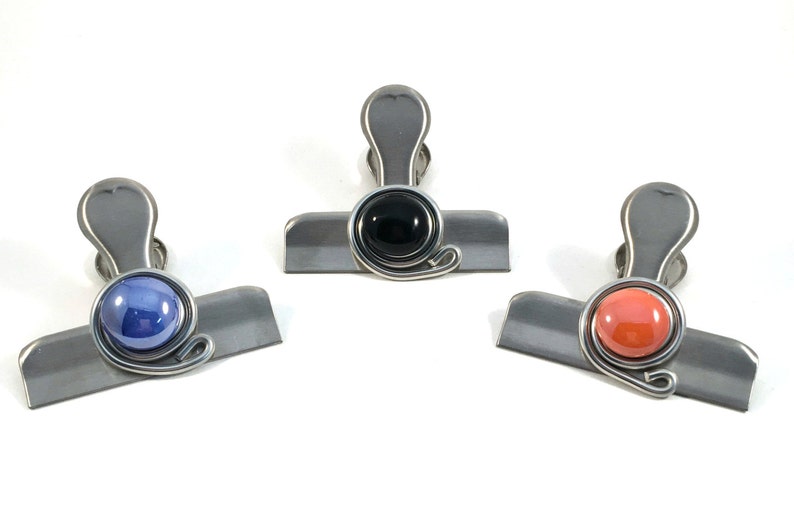 May include: Three metal binder clips with colored accents. One clip has a blue orb, another a black orb, and the third a coral orb. Each clip has a silver-colored body and a decorative wire swirl.