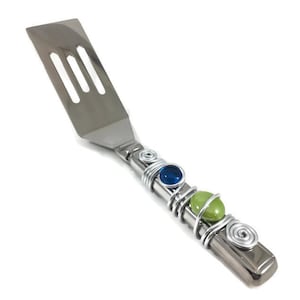 May include: A silver spatula with a blue and green beaded handle. The handle is wrapped in wire and features two blue and two green beads.