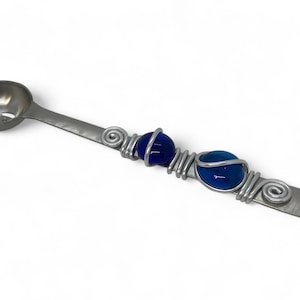 May include: A silver spoon with a small, round bowl and holes for straining. The handle is decorated with three blue glass beads wrapped in silver wire.