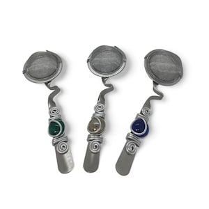 May include: Three silver tea infusers with a mesh strainer and a decorative wire wrapped handle. Each handle has a different colored glass bead: green, brown, and blue.