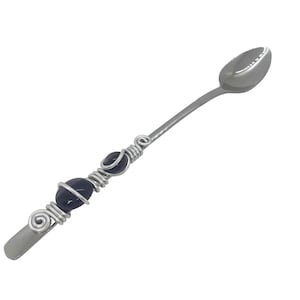 Può includere: Un cucchiaio d'argento con una ciotola arrotondata e un manico decorativo. Il manico è avvolto con filo d'argento e presenta due perle di pietre preziose blu scuro.